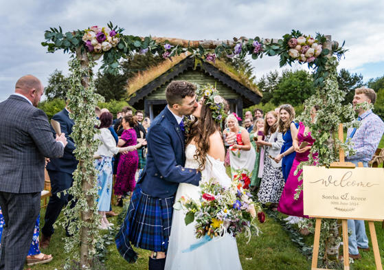 Bride and groom kissing at their wedding ceremony in Scotland, surrounded by family and friends.